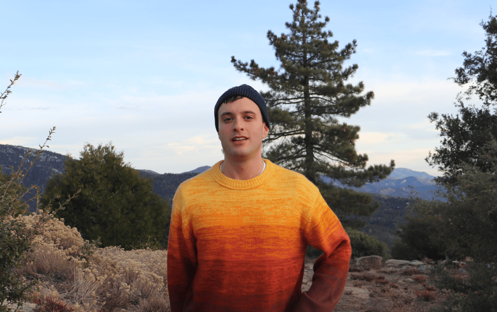 A person standing outdoors in a colorful sweater with an orange gradient, wearing a blue beanie, with a mountainous landscape and trees in the background.